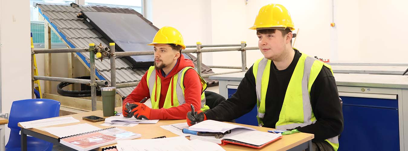 Two men sitting at desk wearing hard hat and hi-vis jacket